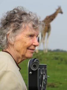 Anne, Bolex camera and giraffes. 2017. Anne Innis Dagg with a 16 mm film camera, and a giraffe in the background (vertical image).