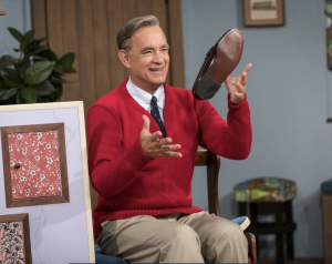 An image of Tom Hanks as Mr. Rogers. He is wearing a red sweater and throwing a shoe in the air.