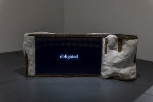 A television screen with a ceramic and wire hand made frame. On the screen is the text "obligated"