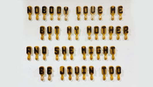 4 rows of hair brushes on a wall. Bristles have been removed to form letters. The brushes accumulated text "Nobody loves me but my mother and she might be jivin too"