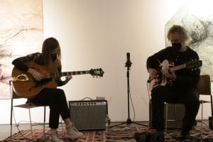 two guitarists improvising together in front of contemporary art hanging on the walls