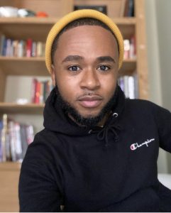 A man staring into the camera wearing a black hooded sweatshirt and a yellow beanie.