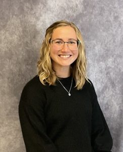 A headshot of a blonde woman wearing glasses and a black shirt.