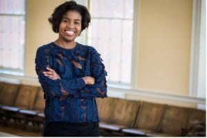 A woman of color in a blue shirt standing in front of a bench with her arms crossed.