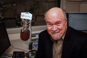 An older man sitting and smiling in front of a microphone.