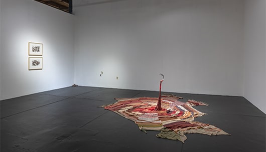 A large woven textile of varying red hues against a black concrete floor. Part of the textile is suspended from the ceiling.