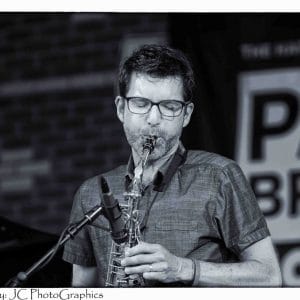 A black and white image of a man in glasses playing a saxophone on a stage.