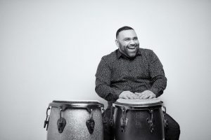 A black and white image of a man smiling and playing drums with his hands.