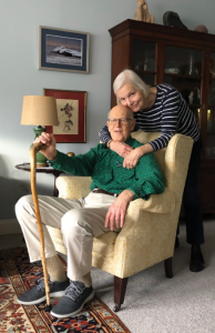 A man sitting in a chair holding a cane with a woman wrapping her arms around him.
