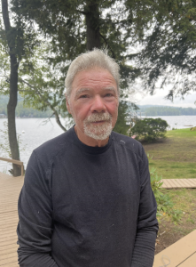 A headshot of a man standing in front of a body of water with trees in the background.